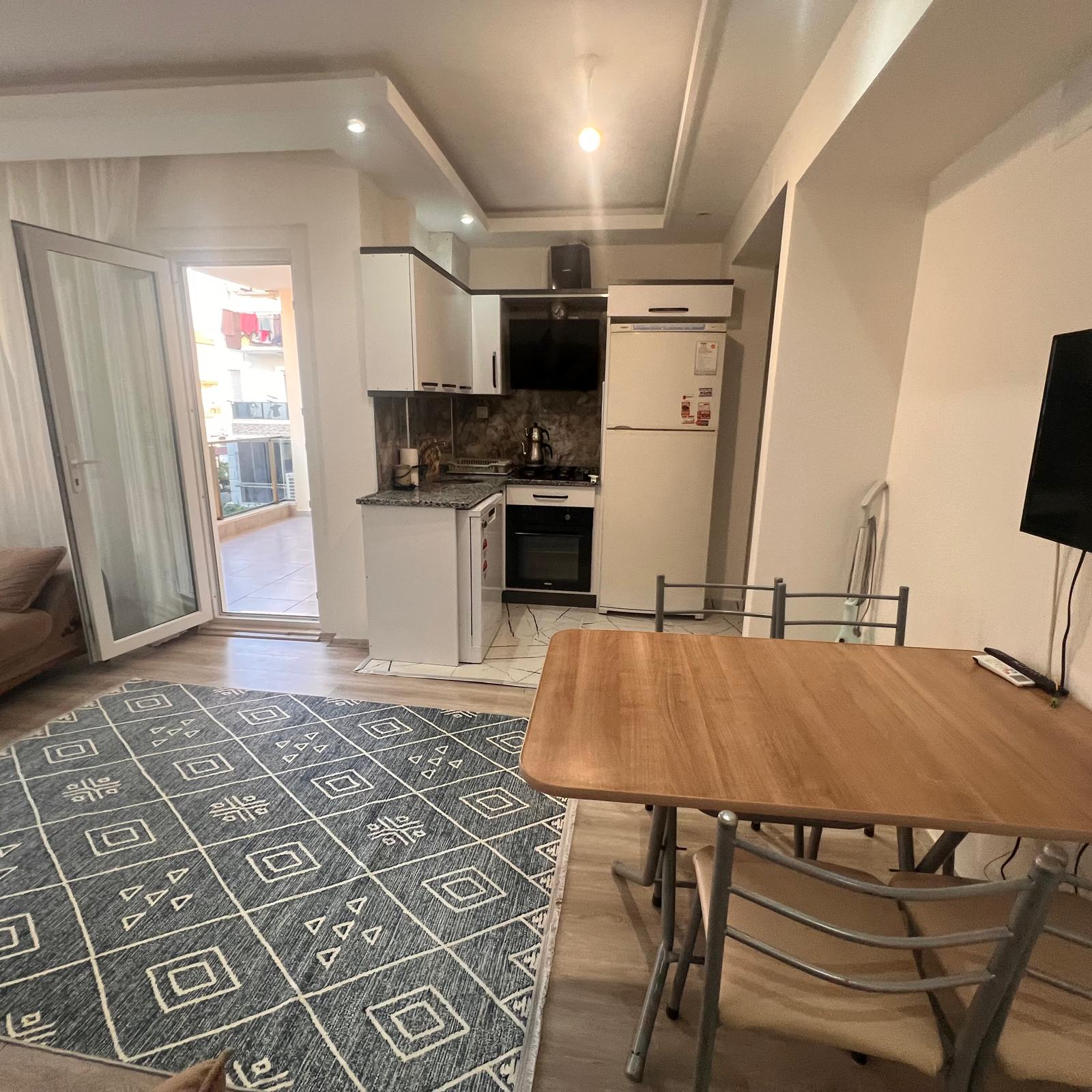 A modern open-plan kitchen and dining area in a short-term rental apartment, photographed with clean and realistic detail. Gleaming white cabinetry with sleek silver handles lines one wall, contrasted by a subtle gray backsplash and a spotless black ceramic cooktop. A compact dining table with a smooth wooden surface is set with two immaculate white plates, clear water glasses, and a small decorative bowl. Natural daylight from an unseen balcony door illuminates the space evenly, reflecting softly off the glossy tiled floor and stainless-steel appliances. The mood is practical, orderly, and reassuring, showcasing full self-catering comfort. Captured at eye level with sharp focus and a slightly wide composition, the image conveys functionality, simplicity, and professional presentation.