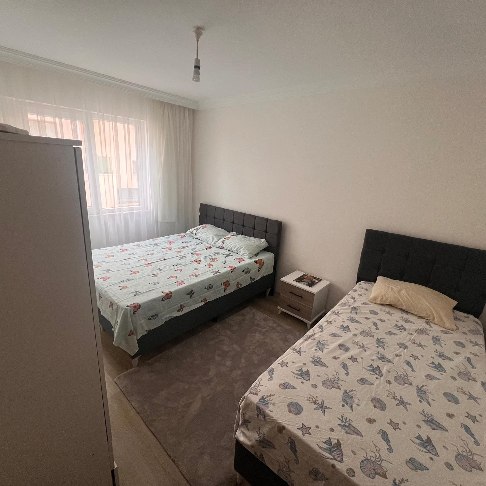 A bright, impeccably clean living room in a modern holiday apartment in Dalaman, photographed in realistic detail. A plush light-gray L-shaped sofa with soft textured fabric surrounds a low natural-wood coffee table with a single neatly folded brochure labeled “Günlük & Haftalık Kiralık.” Large sliding glass doors open to a sunlit balcony with lush green trees and distant hills. Soft Mediterranean daylight fills the space, creating gentle shadows on the glossy tiled floor. The mood is calm, professional, and welcoming, emphasizing comfort and trust. Shot at eye level with a wide lens, sharp focus throughout, and a clean, modern aesthetic that highlights the apartment’s spaciousness and everyday livability.