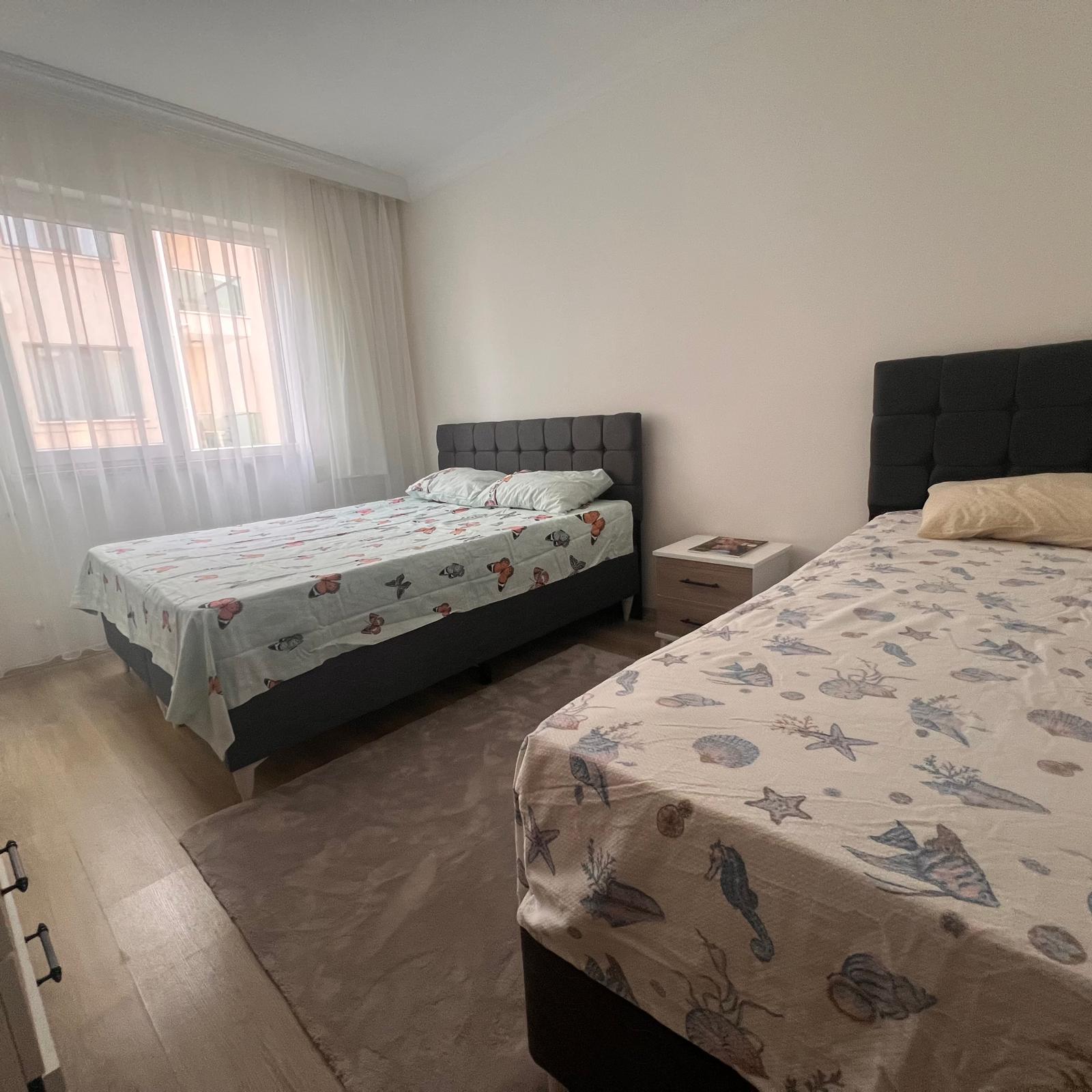 A spacious balcony of a Dalaman holiday apartment overlooking a calm residential neighborhood, rendered in photographic realism. Two empty woven rattan chairs with cream cushions flank a small round glass-topped table, where a simple ceramic plant pot adds a touch of greenery. The sturdy metal railing frames a view of red-tiled roofs, sunlit palm trees, and distant green hills under a clear blue sky. Late-afternoon sunlight bathes the balcony in a warm glow, casting soft linear shadows across the light stone tiles. The scene feels relaxing, private, and safe, ideal for quiet daily or weekly stays. Shot from a slightly elevated corner angle, with balanced composition and moderate depth of field, emphasizing outdoor comfort and Mediterranean atmosphere.