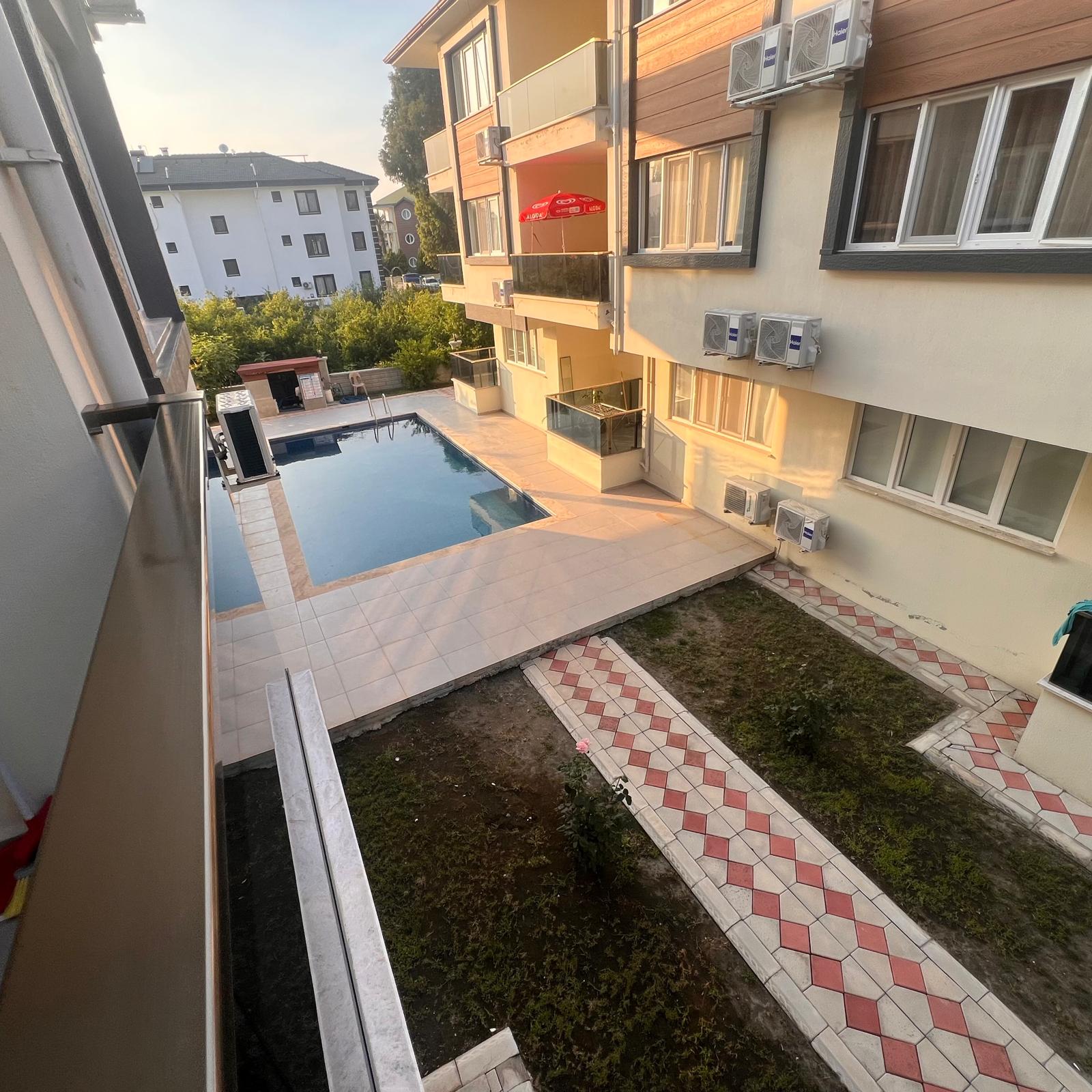 A cozy, sunlit bedroom in a tourism-certified rental apartment in Dalaman, captured in photographic realism. A queen-sized bed with crisp white linens, perfectly fluffed pillows, and a neatly folded beige throw blanket sits against a light taupe wall, with a small, minimalist wooden nightstand beside it. On the nightstand, a subtle sign reads “Rahatınız Garantimiz.” Soft golden afternoon light streams in from a large window with sheer white curtains, creating delicate patterns on the polished tile floor. The atmosphere is serene, hygienic, and inviting. Framed with a slightly elevated angle and balanced composition, the image emphasizes restfulness, quality, and professional hospitality standards.