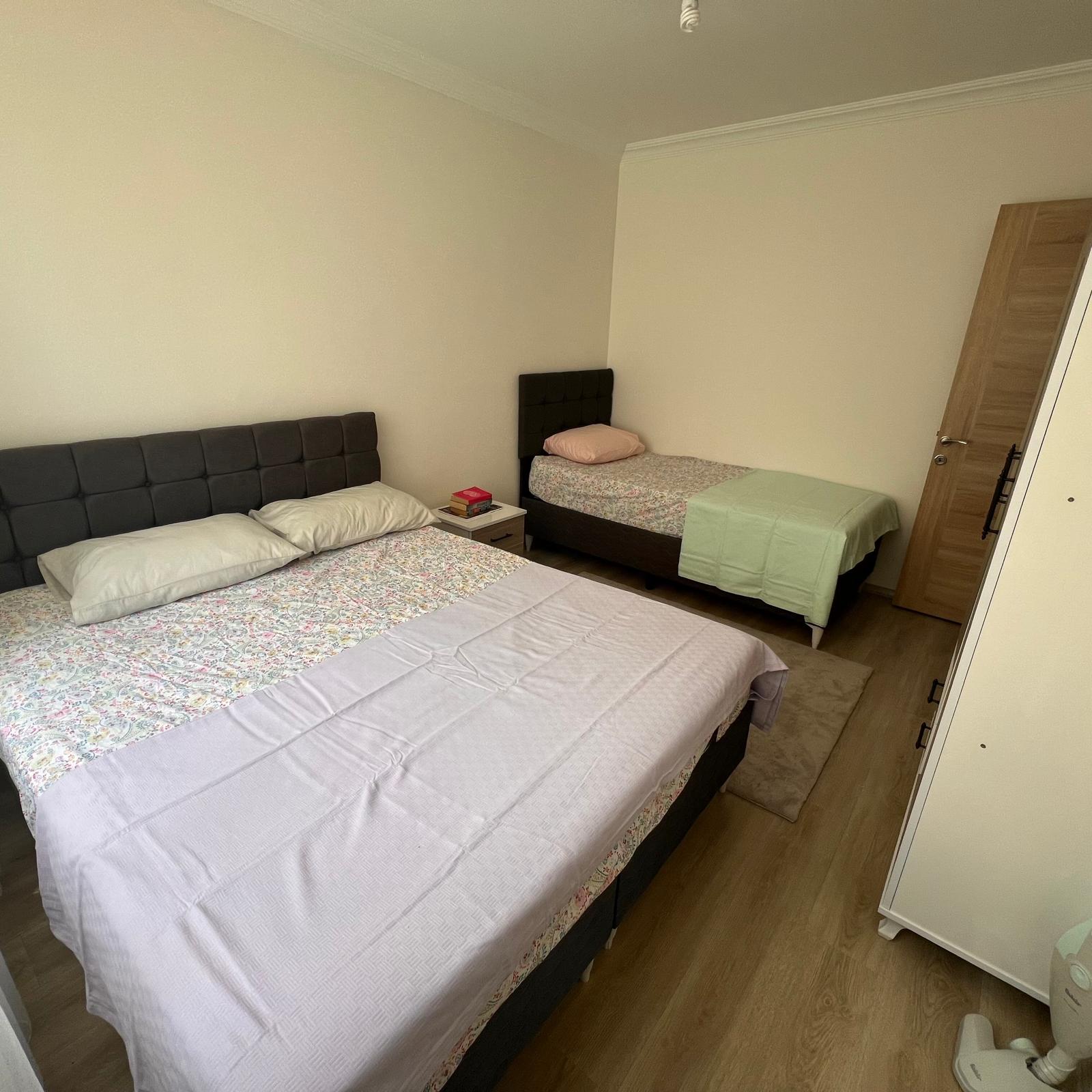 The exterior façade of Yiğit Apart-Dalaman, a well-maintained multi-story apartment building suitable for tourism rentals, captured in crisp photographic realism. The structure features clean white and light-beige walls, symmetrical balconies with sturdy metal railings, and large glass doors that suggest bright interiors. A simple yet clear building sign reads “Yiğit Apart-Dalaman – Turizm Belgeli.” The paved entrance area is spotless, with a few neatly arranged potted shrubs adding greenery. Soft early-evening light creates a gentle, welcoming glow on the façade, with the sky transitioning to a calm pastel blue. Shot from a slightly low, wide-angle perspective, the composition highlights reliability, professionalism, and a sense of arrival for daily and weekly guests.